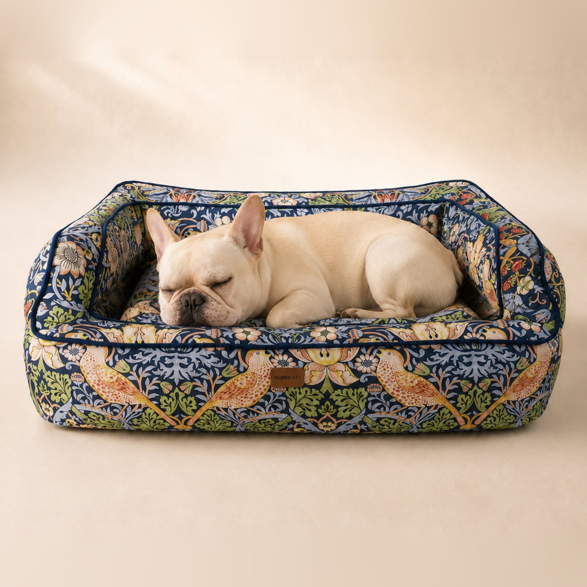 Cream French Bulldog sleeping in a Morris & Co Strawberry Thief pet sofa bed on a warm neutral background