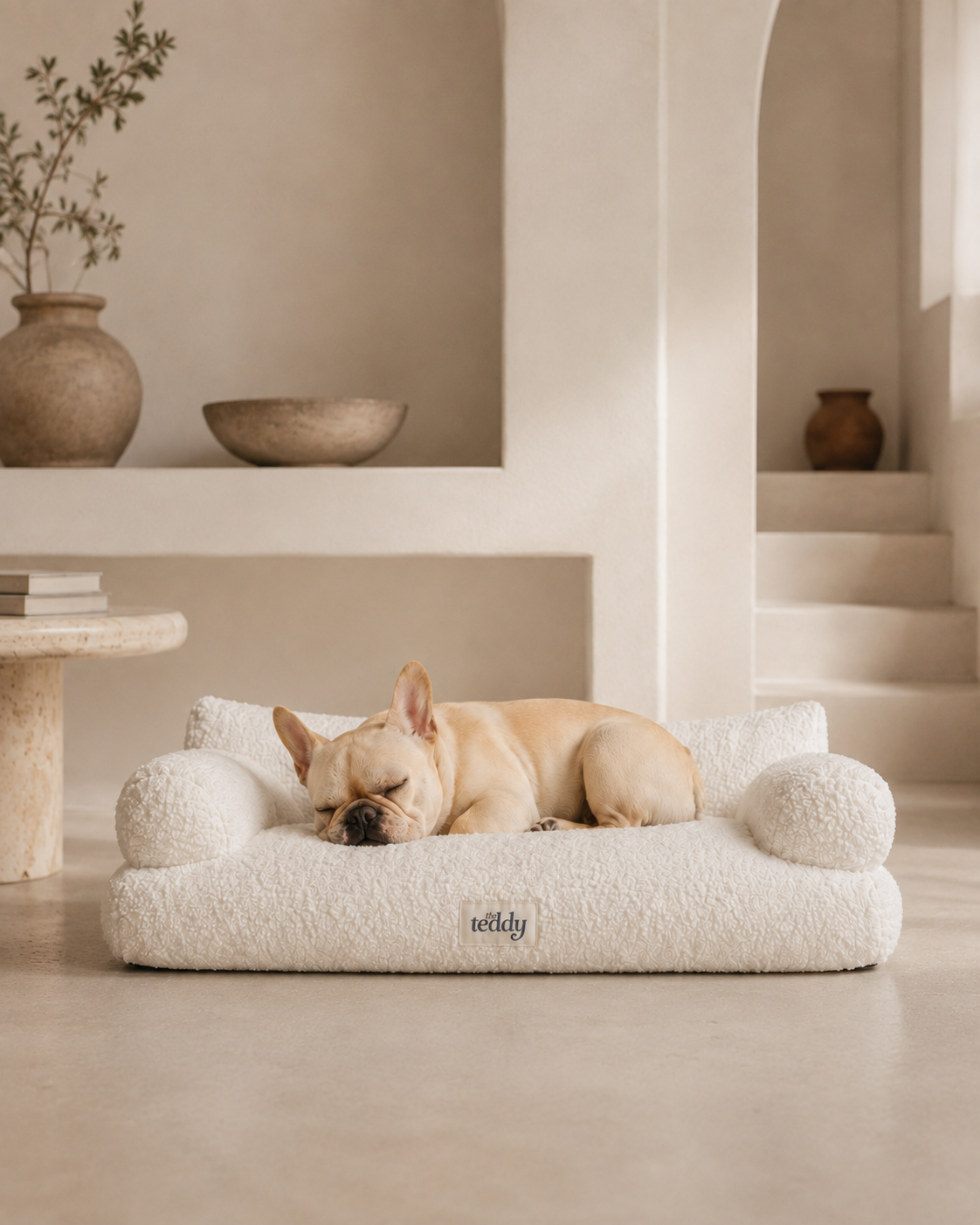 A light tan French Bulldog sleeps peacefully on a fluffy cream boucle dog bed with built-in ball pillows, set in a minimalist, warm-toned modern interior.