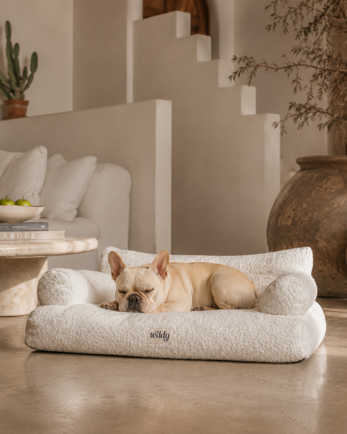 A light beige French Bulldog rests on a plush, cream-colored textured pet bed with 'wicky' embroidered on the front, set in a modern, warm-toned living room.