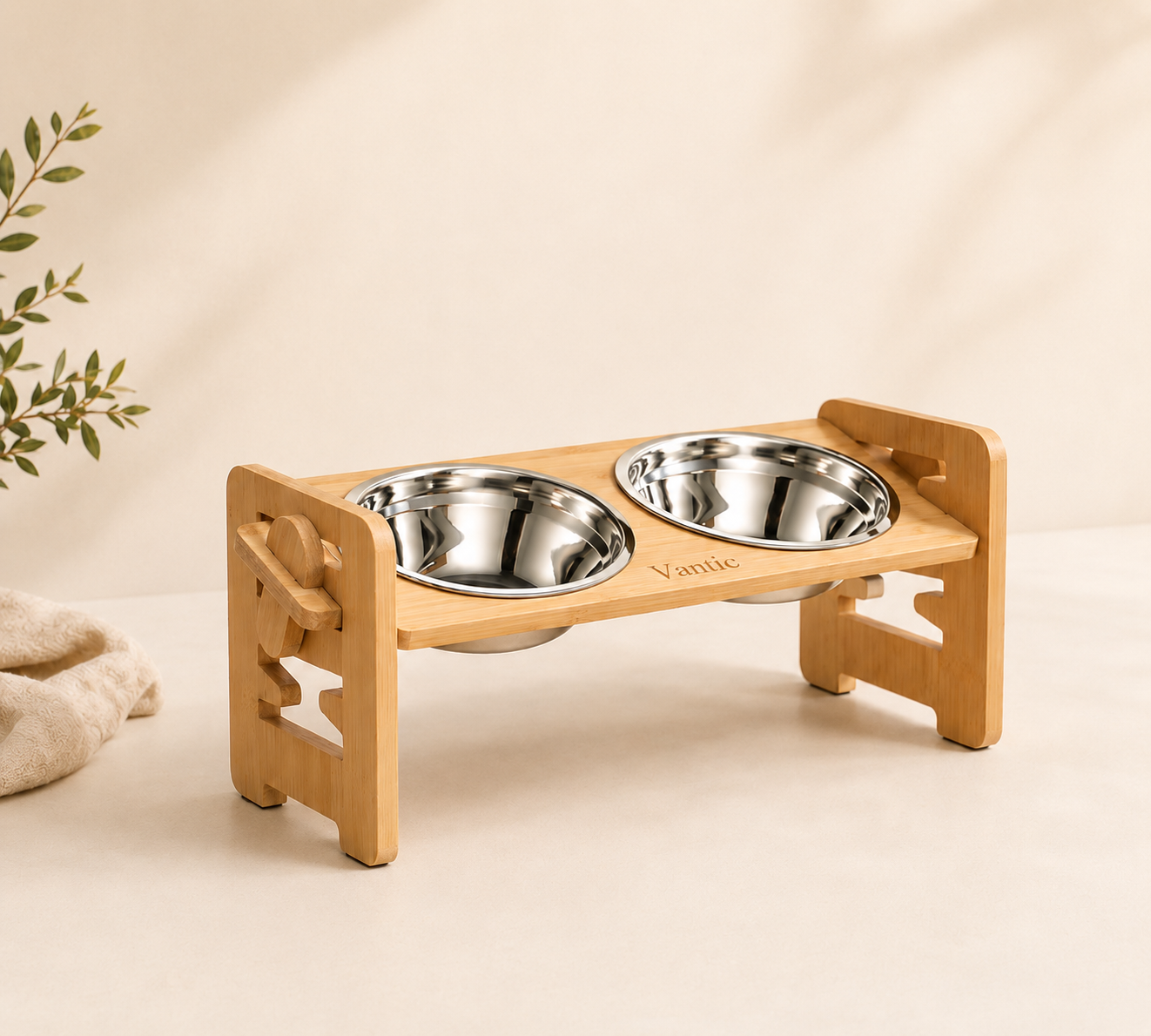 A two-bowl elevated pet feeder stand made of light natural wood with stainless steel bowls, shown on a warm beige background with a small plant and fabric prop at the side.