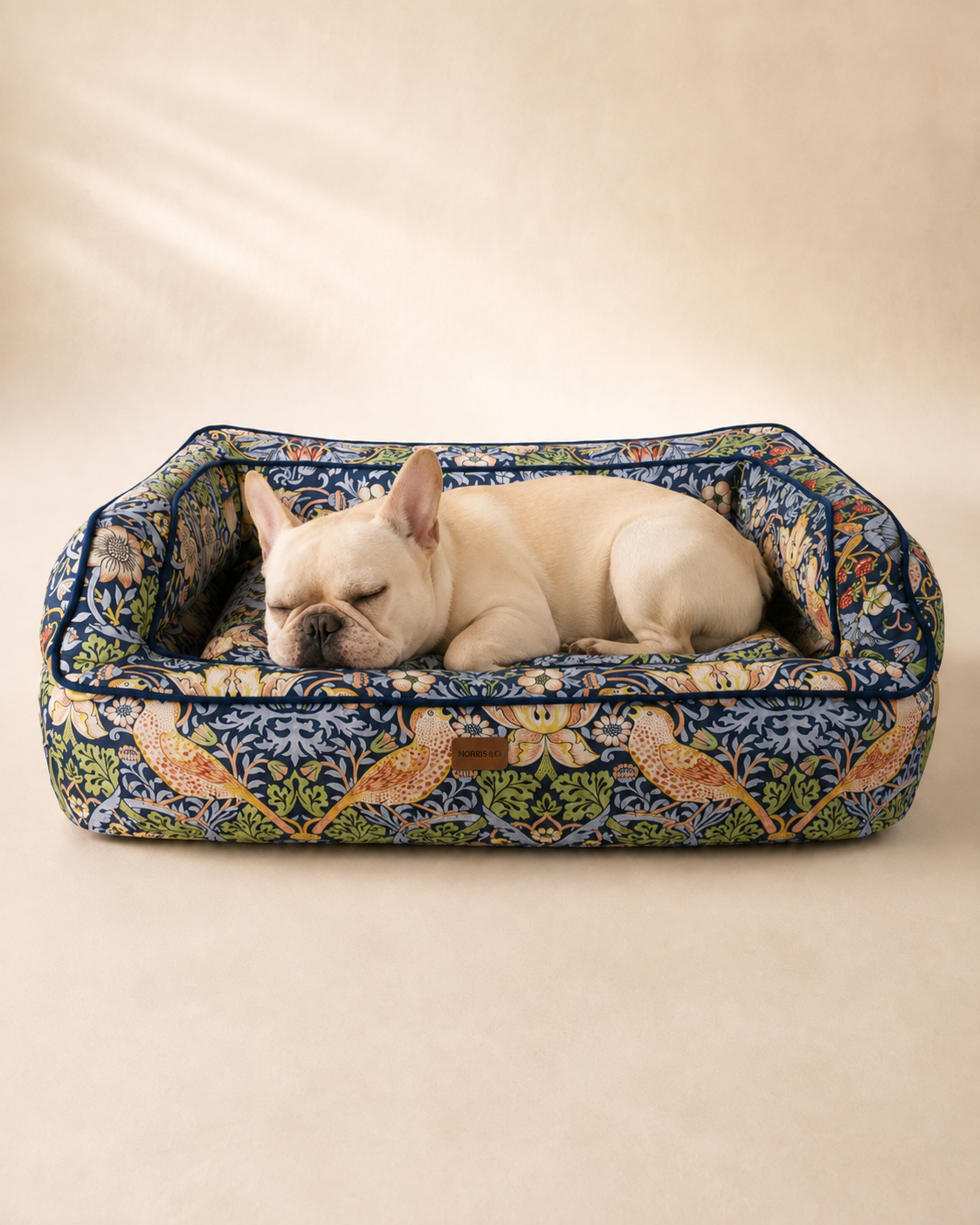 Cream French Bulldog sleeping in a Morris & Co Strawberry Thief pet sofa bed on a warm neutral background